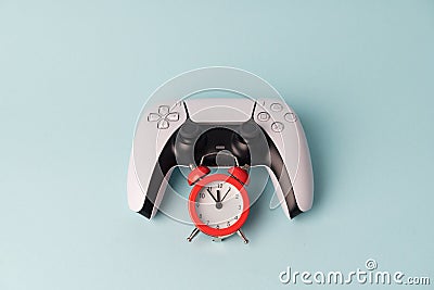 Video game console gamepad on blue background with red alarm clock. Tim to play. Gamer problem in dependance of gaming. Stock Photo