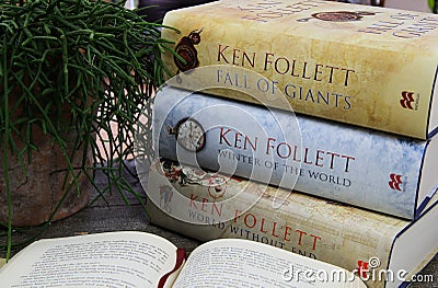View on wood table with open book, stack of Ken Follett novels books and green plant Editorial Stock Photo