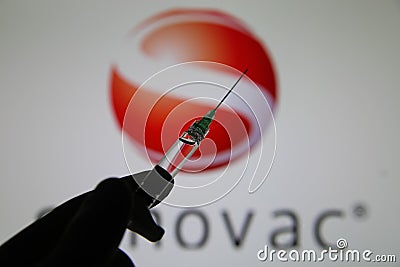 Close up of syringe hold by hand with injection needle and serum, blurred Sinovac logo lettering background Editorial Stock Photo