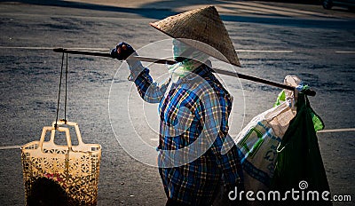 Vietnamese with conical hat carries a yoke on her shoulder along the street. Editorial Stock Photo