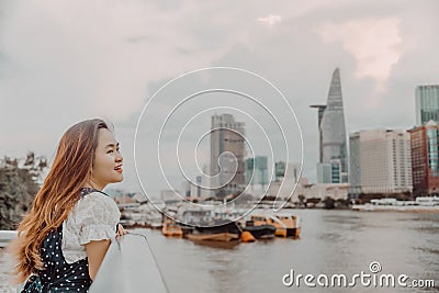 A Vietnamese girl standing by the riverbank admiring the view of Ho Chi Minh city with Bitexco Financial Tower, many buildings and Stock Photo