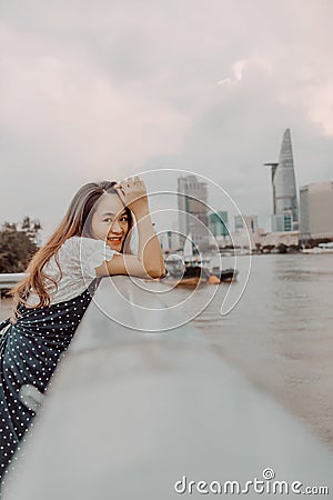 A Vietnamese girl standing by the riverbank admiring the view of Ho Chi Minh city with Bitexco Financial Tower, many buildings and Stock Photo