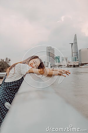 A Vietnamese girl standing by the riverbank admiring the view of Ho Chi Minh city with Bitexco Financial Tower, many buildings and Stock Photo