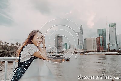 A Vietnamese girl standing by the riverbank admiring the view of Ho Chi Minh city with Bitexco Financial Tower, many buildings and Stock Photo