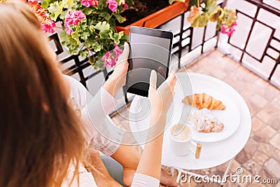 View from above tablet in hands of girl in pajama sitting surround flowers on balcony, having breakfast in the morning. Stock Photo
