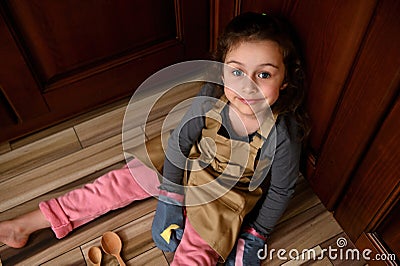 Top view cute child girl, little baker confectioner in a beige chef apron and kitchen mittens sweetly smiling to camera Stock Photo