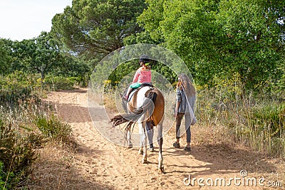 View from behind of little child riding a horse next to her mother walking on track Stock Photo