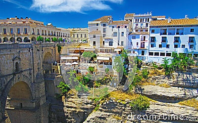 View beyond medieval stone bridge on ancient village on cliff top with cliffside restaurant, blue sky - Ronda, Spain Stock Photo