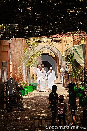 View beyond shade on two muslim men in white robes walking in sunshine alleyway, bright arches background, blurred playing kids Editorial Stock Photo
