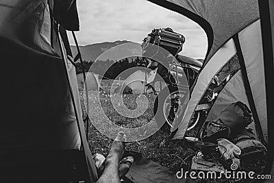 View from camp tent door. Motorcycle adventure bike on the background of green grass, forest and mountains under clouds sky, Stock Photo