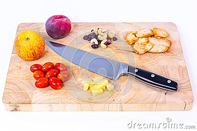 View of a cutting wooden board, an apple, a peach, tomatoes, pieces of pineapple, chicken, raisins and peanuts with a knife Stock Photo