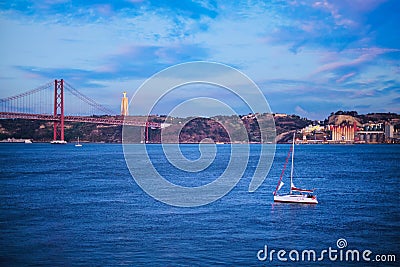 View of 25 de Abril Bridge over Tagus river, Christ the King statue and yacht boat in evening twilight. Lisbon, Portugal Editorial Stock Photo