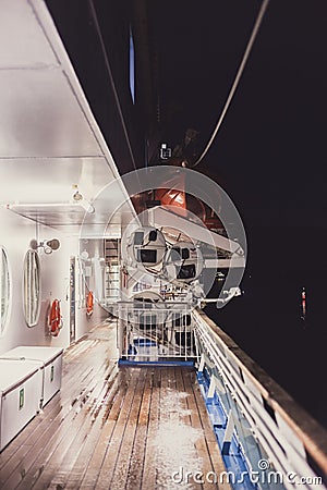 View of the empty ship deck at night with life boats and a lighthouse seen in the water Stock Photo