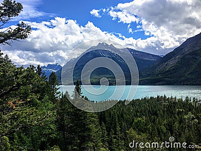 A view of a majestic turquoise lake surrounded by vast green evergreen forests and mountains on a sunny day with blue sky. Stock Photo