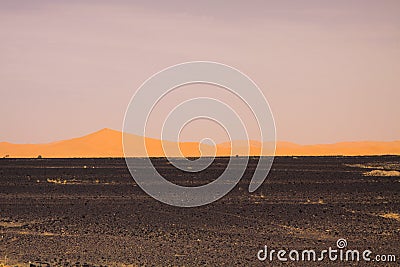 View over endless burned black flat waste stony land on golden sand dunes and blurred gloomy sky, Erg Chebbi, Morocco Stock Photo