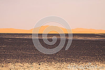 View over endless burned black flat waste stony land on golden sand dunes and blurred gloomy sky, Erg Chebbi, Morocco Stock Photo