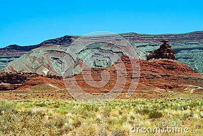 View over plain with green dry grass tufts on red and grey sandstone hill formation butte against blue sky - Monument Valley Stock Photo