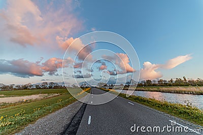 View of a paved street at sunset with grassland, spring flowers and water on both sides, with pink clouds in the air, against a br Stock Photo