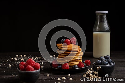 View of plate with stacked pancakes, blueberries, raspberries and bottle of milk, on table and dark background with white flowers Stock Photo