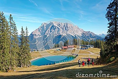 View of rugged Mountain Zugspitze on a brisk sunny day with tourists walking toward a blue pond at the foothills Stock Photo