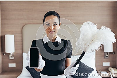 View of smiling maid in white gloves holding duster and smartphone with blank screen in hotel room Stock Photo