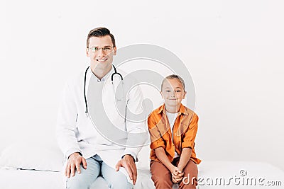 View of smiling pediatrist in white coat and child sitting on hospital bed in clinic Stock Photo