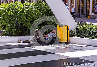 View of tired man with yellow bag sleeping on floor near green bushes on walking street. Miami. USA Editorial Stock Photo