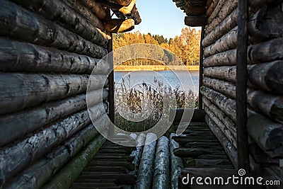 View to the tranquil blue lake, orange autumn trees and reeds through the narrow lane between two old log huts Stock Photo