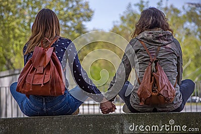 View of two girlfriends girls sitting on a wall and looking at the landscape hand in hand and lovingly Stock Photo
