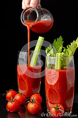 View of two glasses with Bloody Mary cocktail with celery, woman`s hand serving from a jug, and cherry tomatoes, on black backgro Stock Photo