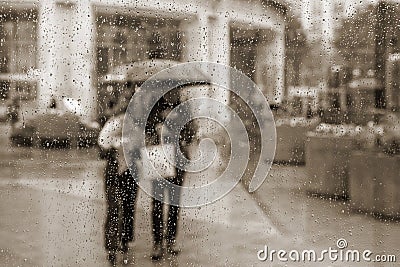 Vintage abstract blurred background. View through glass window with rain drops. People under the umbrellas. Rainy Stock Photo