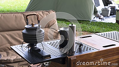 Outdoor black kettle and coffee grinder on camping table with armchair on green lawn in camping area Stock Photo