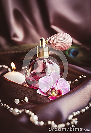 Vintage spray atomizer perfume bottle and pearl jewellery on silky dark pink fabric with peacock feather and candle burning. Stock Photo