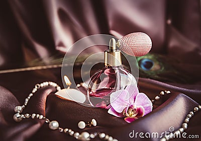 Vintage spray atomizer perfume bottle and pearl jewellery on silky dark pink fabric with peacock feather and candle burning. Stock Photo