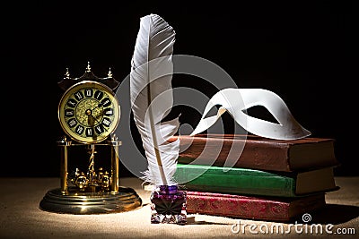 Vintage still life with theater mask on old books near inkstand, feather, old clock against black background under beam of light. Stock Photo