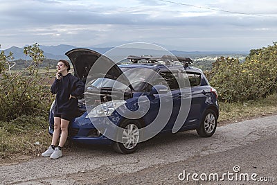 A visibly upset asian girl calls a towing service due to car trouble. Stranded at a remote rural road. A blue hatchback Stock Photo