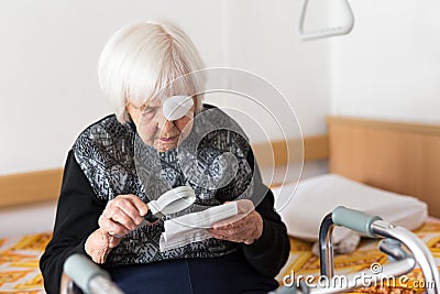 Visually impaired elderly 95 years old woman sitting at the bad trying to read with magnifying glass. Stock Photo