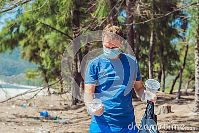 Volunteer man in face mask pick up garbage pollute beach near sea, hold look at discarded disposable plastic cups Stock Photo