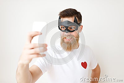 Waist up portrait of cheerful bearded male model poses at smart phone camera, wears batman mask and casual t shirt Stock Photo