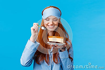 Waist-up portrait cheerful and delighted young caucasian redhead woman in pyjama and sleep mask, holding slice cake and Stock Photo