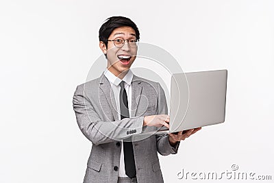 Waist-up portrait of excited, happy smiling asian businessman in grey suit, standing white background, typing at laptop Stock Photo