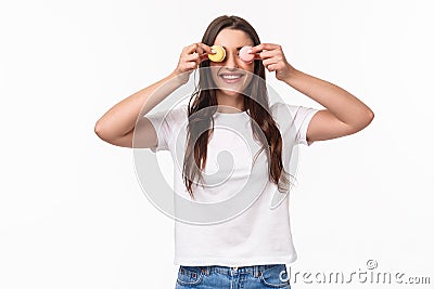 Waist-up portrait of funny and cute, adorable young woman holding two desserts macarons over eyes and smiling, playing Stock Photo