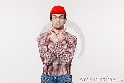 Waist-up portrait funny indecisive guy in glasses and red beanie dont know what choose, pointing sideways at left and Stock Photo