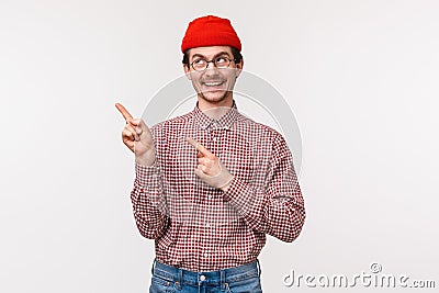 Waist-up portrait of happy smiling geeky guy in glasses and red beanie, pointing and looking upper left corner with Stock Photo
