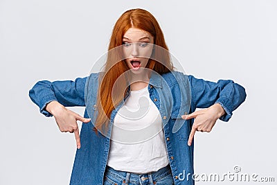 Waist-up portrait impressed, curious and astonished redhead cheerful girl in denim shirt, drop jaw, looking and pointing Stock Photo