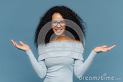 Waist up shot of happy young crisp Afro American woman raises both hands, pretends holding something, wears blue sweater, has Stock Photo