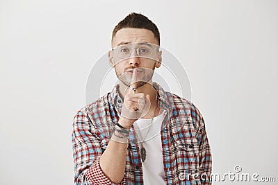 Waist-up shot of kind friendly man in glasses, saying shush with slight smile, holding index finger over mouth, asking Stock Photo