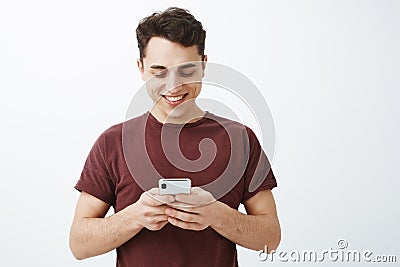 Waist-up shot of positive friendly handsome man in red t-shirt, holding smartphone and smiling joyfully at screen while Stock Photo