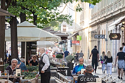 Waiter, a server in uniform, working at the terrace of a cafe serving food to clients wearing a respiratory face mask Editorial Stock Photo