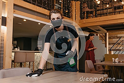 Waitress in face mask holding bottle with sanitizer and cleaning table with rag Stock Photo